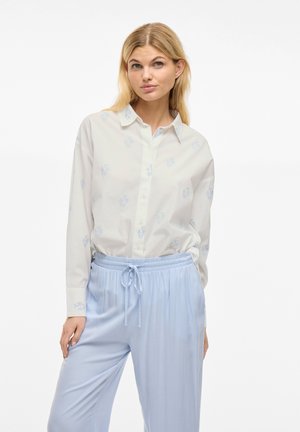 Camicia bianca con motivi di ancore azzurri, abbinata a pantaloni azzurri con cordoncino. Entrambi i tessuti sembrano morbidi e casual.
