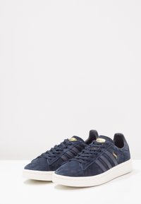 Tmavě modré semišové tenisky s třemi texturovanými pruhy, zlatými logy, plochými tkaničkami a bílou gumovou podrážkou. Klasický nízký střih.