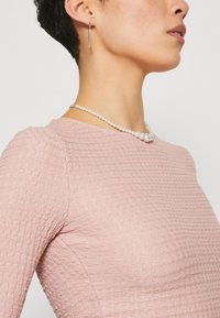 Gewebtes rosa Texturtop mit rundem Ausschnitt, kombiniert mit einer Perlenkette und einem silbernen, länglichen Ohrring, der ein Nahaufnahme-Profil zeigt.