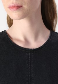 Black denim garment with a rounded neckline, featuring minimal stitching and a smooth texture, exposing the shoulder area.