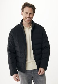 Usměvavý muž v černé péřové bundě přes béžové polo tričko a šedé džíny, stojící před jednoduchým bílým pozadím.