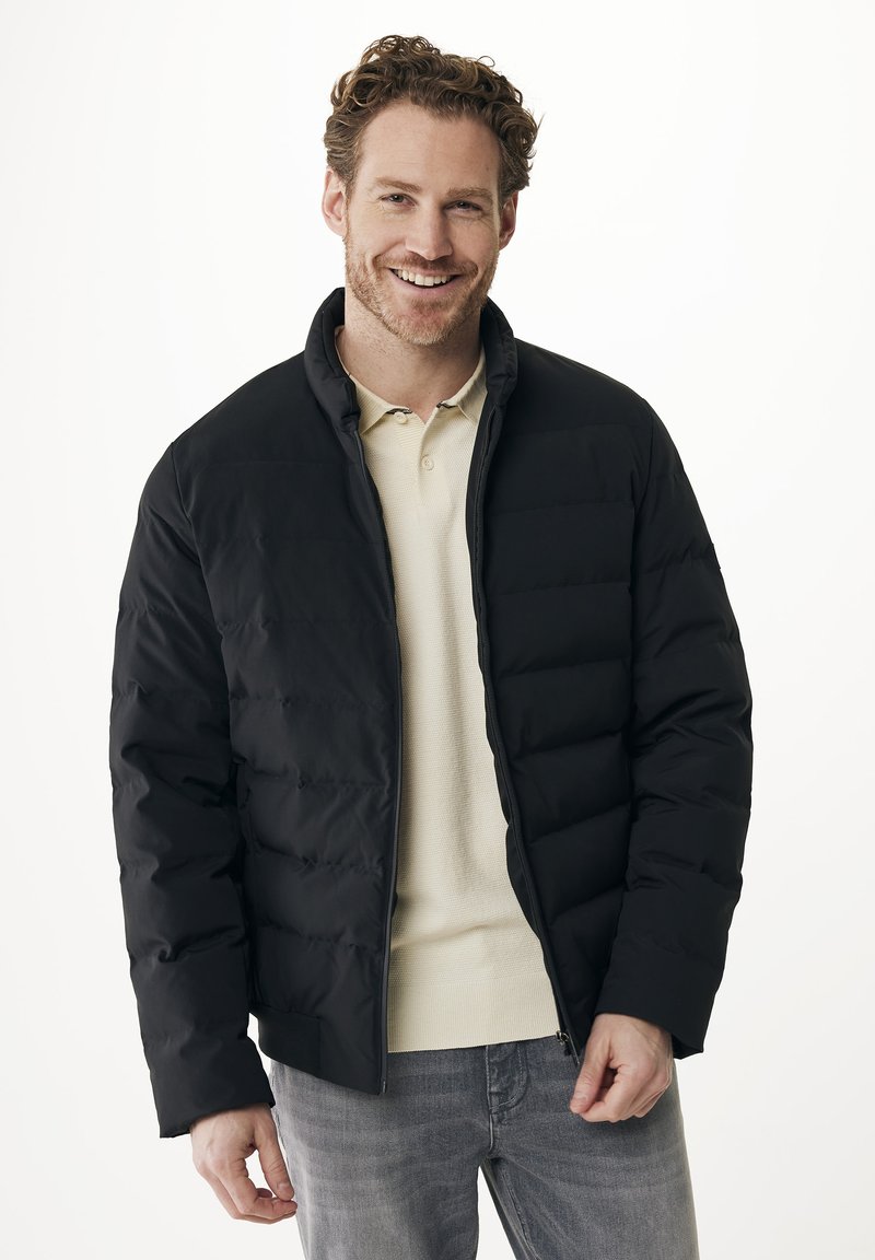 Usměvavý muž v černé péřové bundě přes béžové polo tričko a šedé džíny, stojící před jednoduchým bílým pozadím.