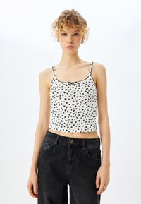 Hvid crop top med sort blomstermønster, tynde stropper og en bølget kant. Kombineret med højtaljede sorte denimjeans.
