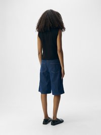 Top negro sin mangas, pantalones cortos de mezclilla en azul oscuro con bolsillos traseros visibles y costuras claras. Zapatos planos negros completan el atuendo.