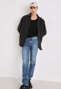 Zwarte oversized shirt, zwarte tanktop, lichtblauwe spijkerbroek en zwarte schoenen. De persoon draagt een beige pet met een logo.