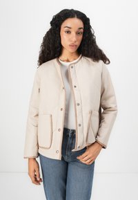 Beige kviltad jacka med tryckknappar, kontrasterande kantband och två framfickor. Bärs över en ljus topp, tillsammans med blåjeans.