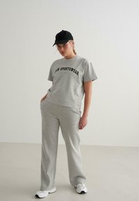 Grå sportsoutfit bestående af en kortærmet skjorte med sort tekst, matchende sweatpants og en sort kasket, sat mod en lys baggrund.