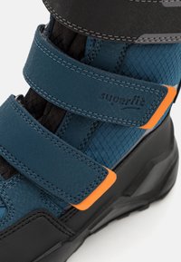 Blue and black ankle-height shoe featuring textured fabric, leather overlays, Velcro straps, and an orange accent near the sole.
