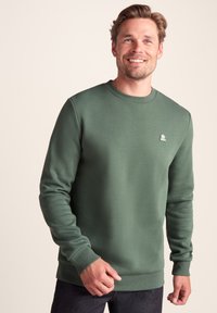 Smiling man wearing a green long-sleeve sweatshirt with ribbed cuffs and a small chest logo, posing against a plain beige background.