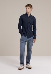 Marineblauwe blouse met knopen en kraag, gecombineerd met lichtblauwe spijkerbroek en witte sneakers. Eenvoudig ontwerp, slim fit en een gladde textuur.