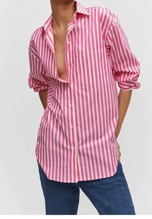 Personne portant une chemise à rayures verticales roses et blanches avec manches retroussées et un jean bleu, main dans la poche, sur fond uni.