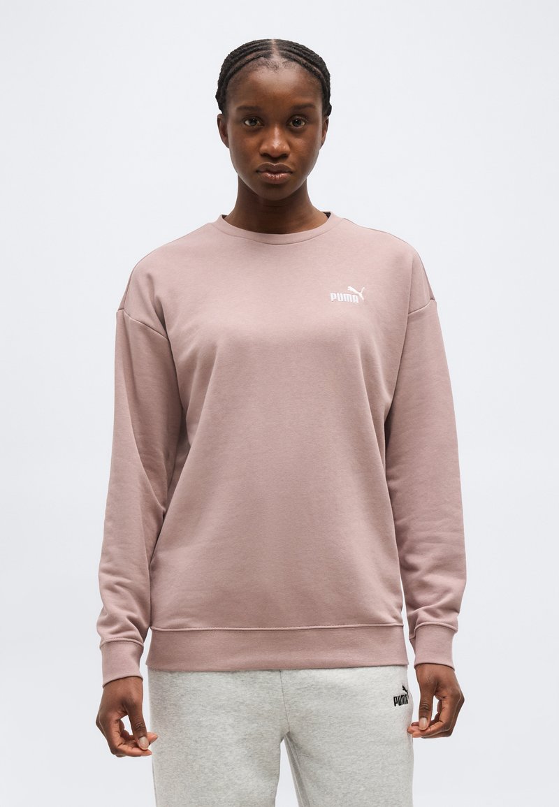 Γυναίκα με πλεξούδες που φοράει μαβιέ φούτερ Puma και ανοιχτό γκρι παντελόνι φόρμας Puma, στέκεται μπροστά σε απλό φόντο.