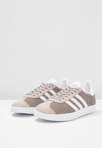 Ljust beige sneakers med vävt textilöverdel, mockadetaljer, vit tre-stripesdesign och gummisula. Har snörning.