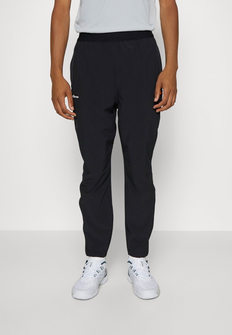 Muž v černých sportovních kalhotách a bílých teniskách stojící na neutrálním, světlém pozadí.