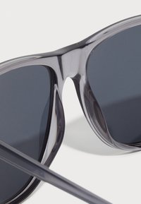 Ochelari de soare cu o ramă gri transparentă, lentile întunecate și o textură netedă. Designul prezintă o formă modernă și temple subțiri.