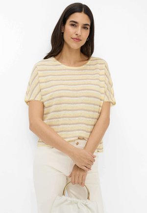 Frau mit langen dunklen Haaren, die ein gelb-weiß gestreiftes kurzärmeliges Oberteil und helle Hosen trägt, hält eine weiße Tasche mit goldenem Ringgriff.