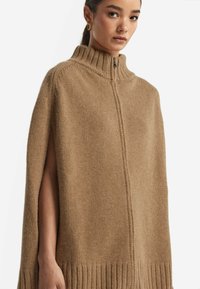 Kamelfärgad stickad cape med hög krage, framdragkedja och ribbad nederkant. Har öppna sidor för enkel rörelse och en strukturerad yta.