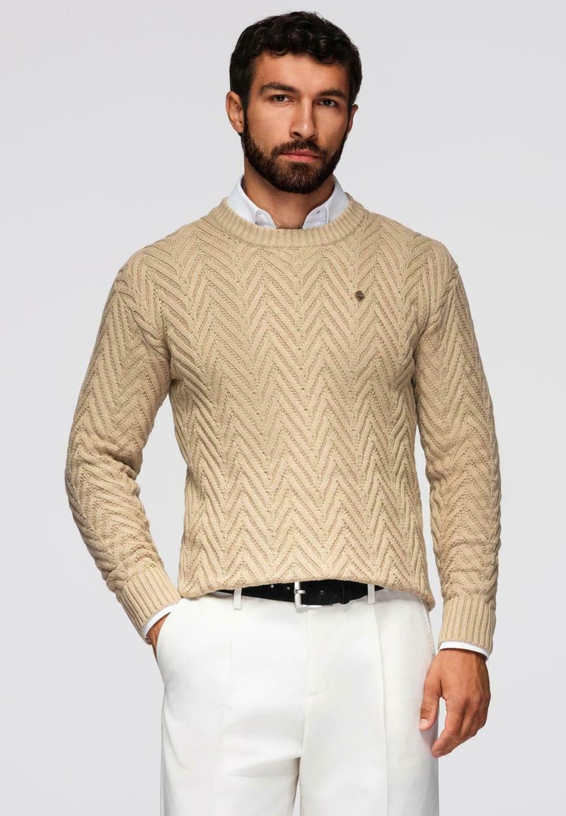 Maglione beige lavorato a maglia con motivo a spina di pesce, scollo rotondo e polsini a coste, abbinato a una camicia bianca e una cintura nera.