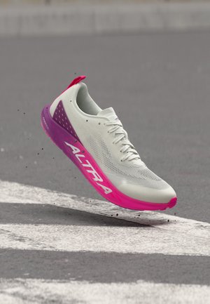 Chaussure de course blanche et violette avec semelle rose et marquage "Altra", capturée en l'air au-dessus d'une route avec des marques blanches.