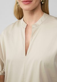 Beige Bluse mit Mandarin-Kragen und einer Schlüsselöffnung. Kurze, geraffte Ärmel. Glattes, leichtes Material. Ohrringe in Goldfarbe.