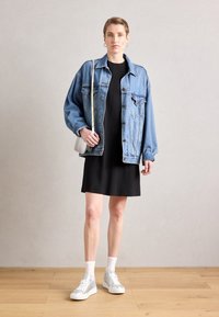 Giacca di jeans con vestibilità over, abito nero, sneaker argento, calzini bianchi e una piccola borsa grigia. Pavimento in legno e sfondo neutro.