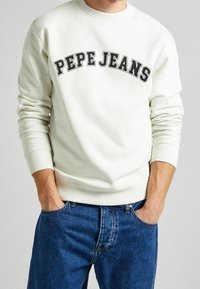 Felpa bianca con "PEPE JEANS" stampato in grassetto nero, polsini e orlo a coste, abbinata a jeans in denim blu.