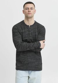 Fekete, hosszú ujjú henley póló texturált mintával, kontrasztos tengerészkék nyakkal és három gombos részlettel. Kényelmesen, keresztbe tett karokkal viselve.
