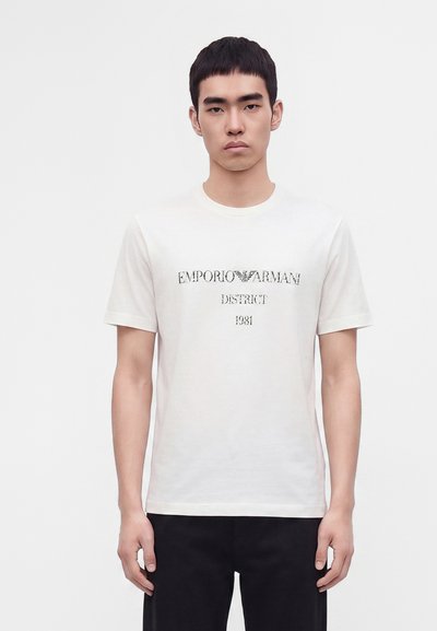 Млад мъж с къса черна коса, носещ бяла тениска Emporio Armani с черен надпис и черен панталон пред обикновен бял фон.