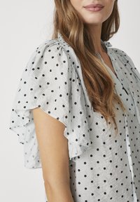 Blusa azzurra chiaro con pois neri, maniche corte a volant e vestibilità comoda. Realizzata in un tessuto morbido e testurizzato.