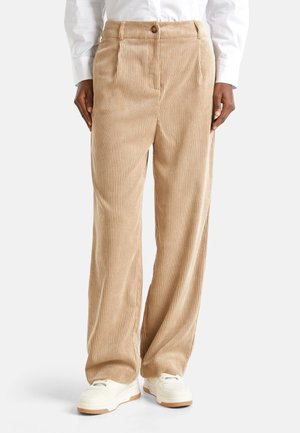 Personne portant un pantalon large en velours côtelé beige, une chemise blanche à manches longues et des baskets blanches, se tenant devant un fond uni.