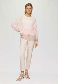 Pull en tricot rose clair avec un motif en chevron, manches larges, et un dégradé vers un ourlet rose plus foncé, associé à un pantalon beige clair ajusté.