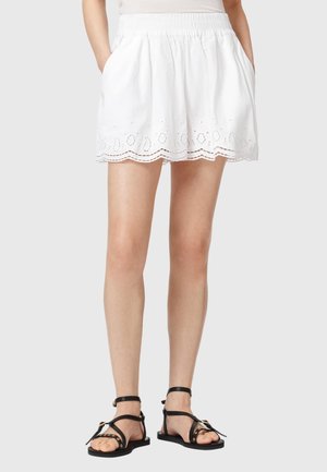 Witte shorts met elastische taille en geborduurde gegolfde zoom, gedragen door een persoon met handen in de zakken, gecombineerd met zwarte platte sandalen met bandjes.