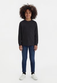 Lapsi, jolla on kiharat hiukset, pukeutunut mustaan pitkähihnaiseen paitaan, sinisiin farkkuihin, mustiin sukkiin ja valkoisiin lenkkareihin, seisoo yksinkertaisella valkoisella taustalla.