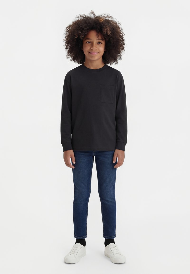 Lapsi, jolla on kiharat hiukset, pukeutunut mustaan pitkähihnaiseen paitaan, sinisiin farkkuihin, mustiin sukkiin ja valkoisiin lenkkareihin, seisoo yksinkertaisella valkoisella taustalla.