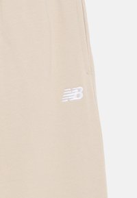 Beige sweatpants with a textured finish, featuring a white embroidered New Balance logo on the thigh and side pockets for functionality.