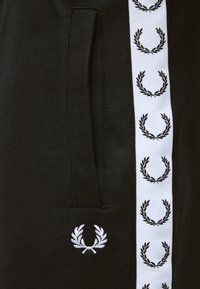 Tissu noir avec une bande verticale blanche comportant des logos répétés de couronne de laurier noire et un petit logo blanc de couronne de laurier sous une poche.