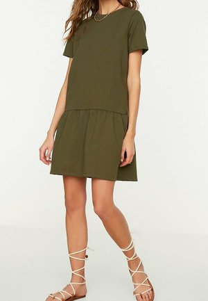 Vrouw draagt een olijfgroene korte mouw jurk met een geplooide rok, gecombineerd met witte platte sandalen met veters.