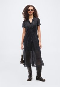 Γυναίκα με σγουρά μαλλιά που φοράει μαύρο φόρεμα με πουά, μαύρες μπότες μέχρι το γόνατο, γυαλιά ηλίου και κρατάει μια μικρή τσάντα με αλυσίδα.