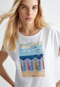 T-shirt bianca con una vivace scena da spiaggia che presenta cabine da spiaggia a righe, mare blu e cielo. Il testo grafico recita "FORTE DEI MARMI."
