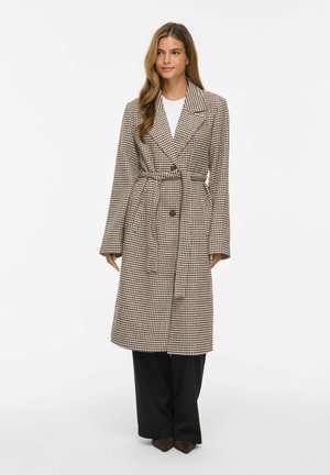Femme portant un long manteau ceinturé à motif pied-de-poule par-dessus un haut blanc et un pantalon noir à jambes larges, debout devant un fond blanc uni.