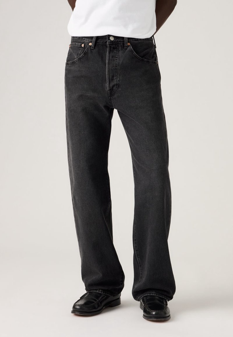 Personne portant un jean noir ample, des chaussures en cuir noires et une chemise blanche sur un fond clair uni.