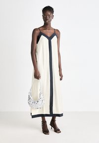 Cremfarbendes, ärmelloses Kleid mit schwarzem Saum und V-Ausschnitt, gerader Silhouette, kombiniert mit einer silbernen, mit Pailletten besetzten Handtasche und schwarzen, hohen Sandalen.