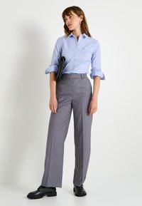 Camisa de botones azul clara, pantalones gris con rayas finas de corte ajustado y un fit relajado, y zapatos negros combinados con un bolso de mano negro.