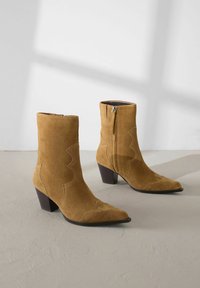 Pari beige suède ankle boots dengan ujung runcing, tumit blok, ritsleting samping, dan jahitan dekoratif di atas permukaan abu-abu muda.
