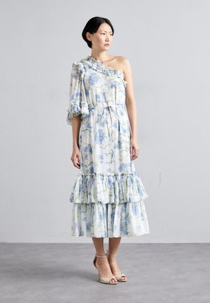 Robe florale à une épaule en tissu léger avec des motifs de plantes bleues et vertes. Présente une manche à volants et une jupe à volants. Accessoirisée avec des talons beiges.