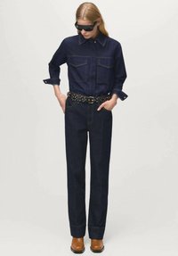 Camicia in denim scuro con due tasche sul petto, abbinata a pantaloni in denim scuro a vita alta. Indossata con una cintura borchiata e stivali marroni.