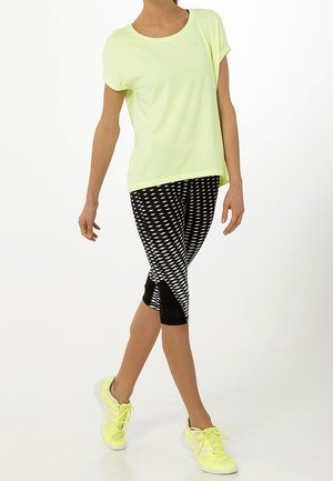 Kvinde iført lysegul kortærmet top, sorte og hvide mønstrede korte leggings og neongule løbesko, stående med krydsede ben.