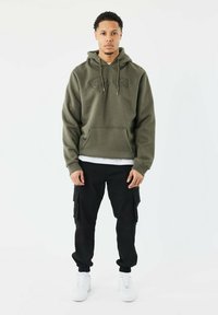 Haut à capuche olive avec poche avant et broderie "Prolific Paris", associé à un pantalon cargo noir et des baskets blanches. Tenue simple et décontractée.