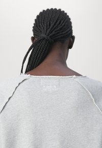 Sweatshirt gris avec un col brut, une texture douce et un logo visible à l'arrière. Le design comporte des manches raglan et une coupe décontractée.