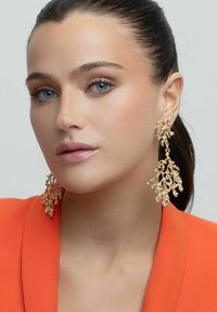 Guldörhängen i korallform med intrikata detaljer och en texturerad yta, bärs tillsammans med en orange blazer. Bakgrunden är neutral.